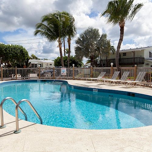 The image shows an outdoor swimming pool surrounded by lounge chairs, palm trees, and houses, with a partly cloudy sky above.