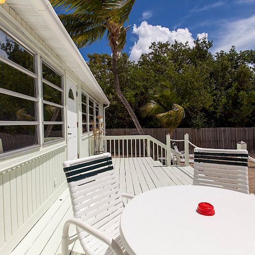 A deck with white chairs and a round table, adjacent to a house with large windows, overlooks a tropical garden and a palm tree.