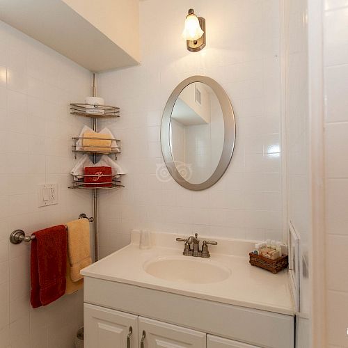 A small bathroom with a round mirror, sink, wall-mounted shelves with towels, a light fixture above the mirror, and a towel rack with towels.