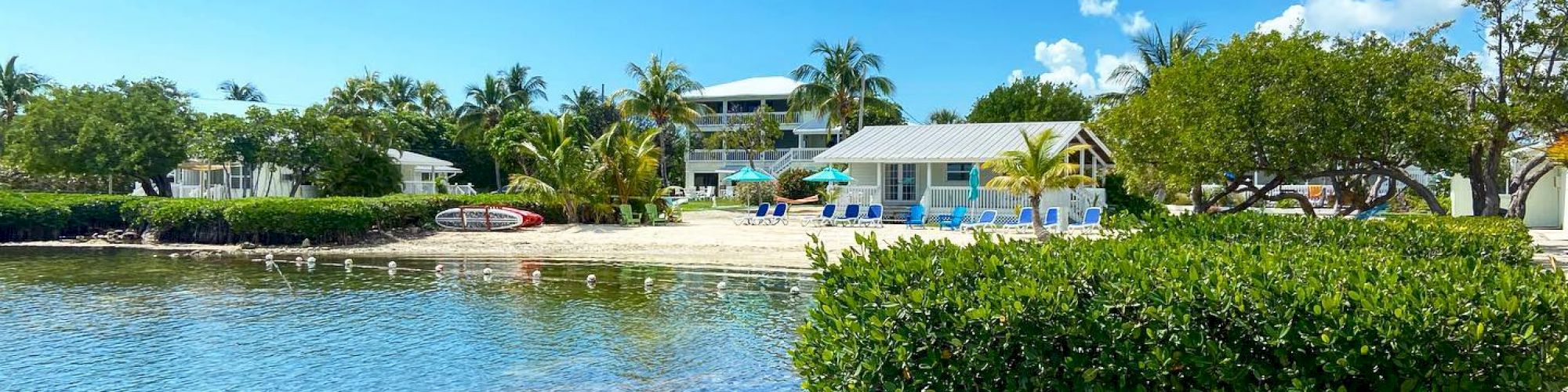 A beautiful beachside scene featuring clear water, lush greenery, palm trees, and quaint buildings under a bright blue sky with scattered clouds.