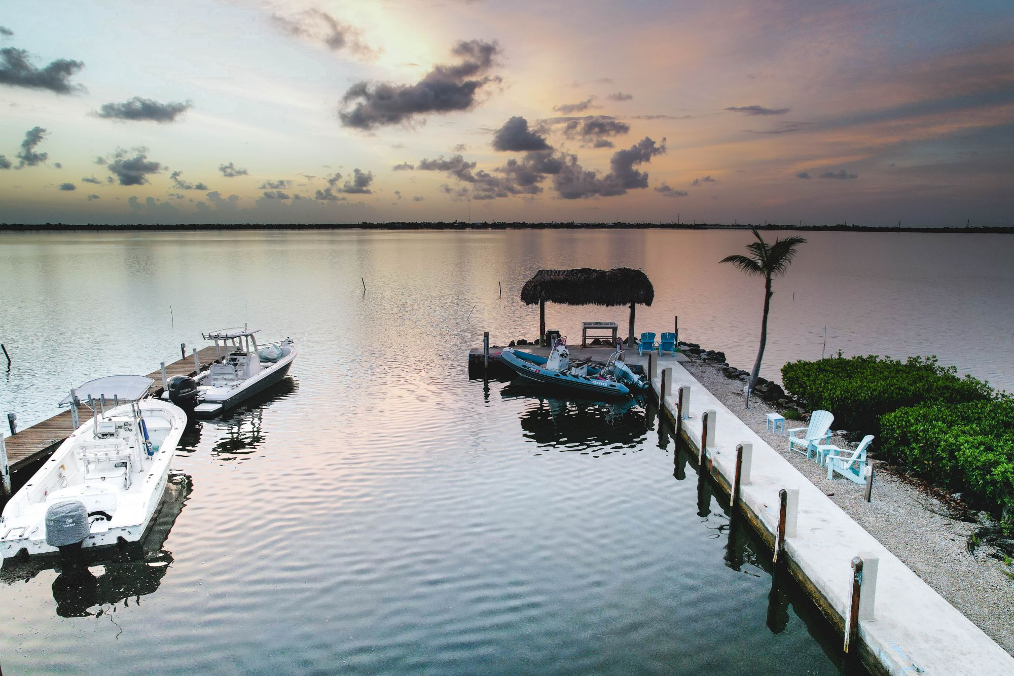 A serene waterfront scene at sunset with two boats docked, a thatched-roof structure, palm trees, and chairs on a pier.