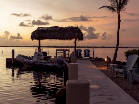 A serene dock at sunset with a thatched roof gazebo, boats, and a lone palm tree, all reflecting serene water.