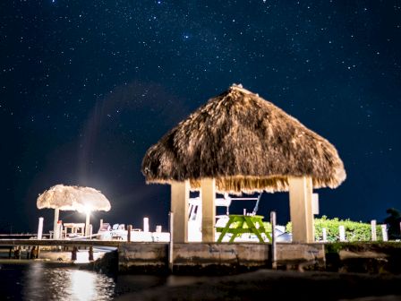 The image shows two thatched-roof structures by a waterfront, illuminated at night under a starry sky.