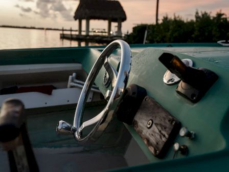 A close-up of a boat's steering wheel and controls against a sunset, with a thatched-roof structure and calm water in the background.