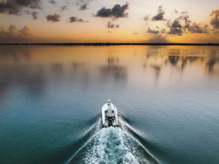 A boat is sailing on calm water towards a scenic sunset, leaving a trail of ripples behind it, under a sky dotted with clouds.