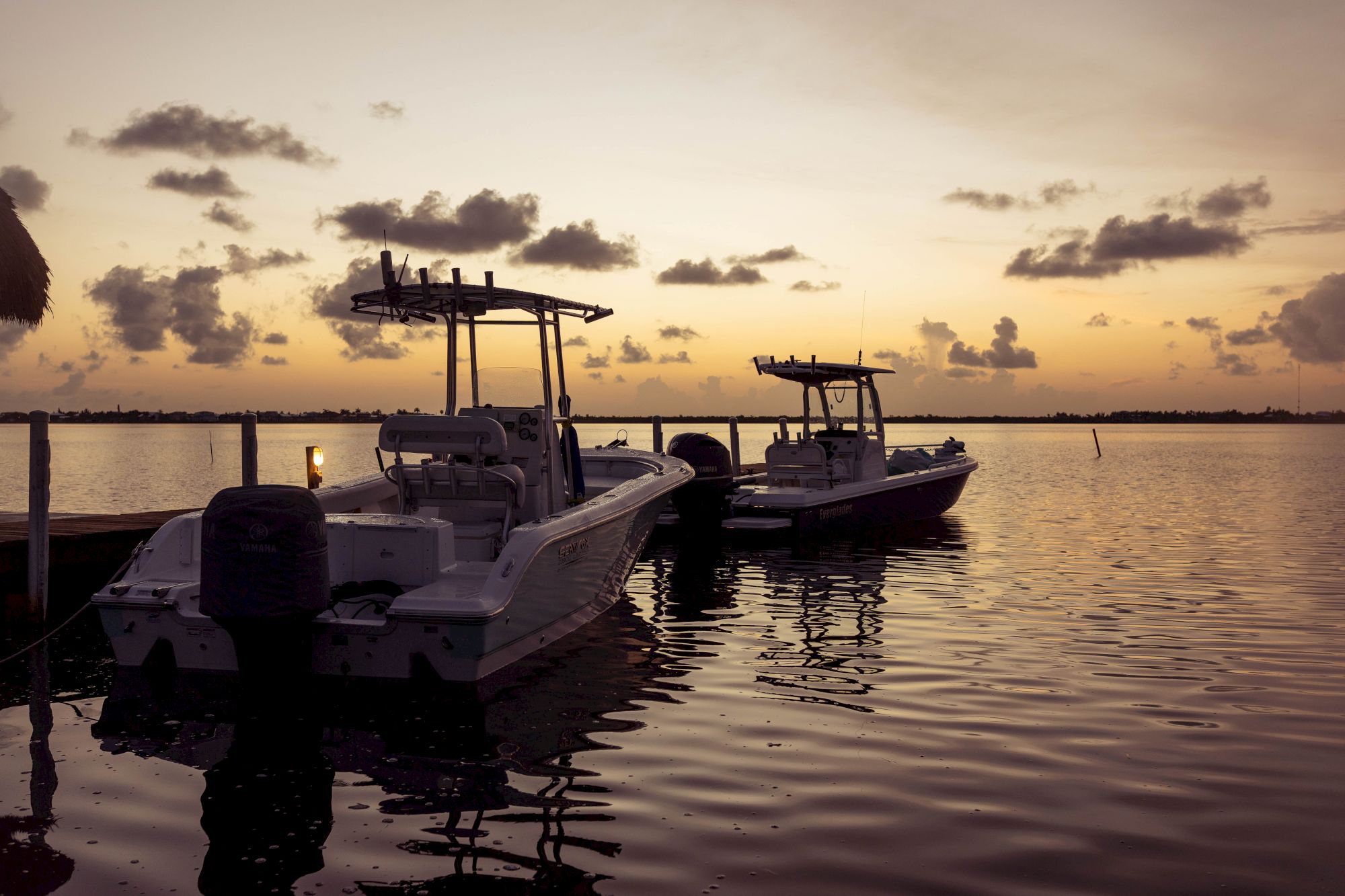 Two boats are docked on calm waters under a golden sunset sky, with silhouetted clouds and a serene horizon in the background.