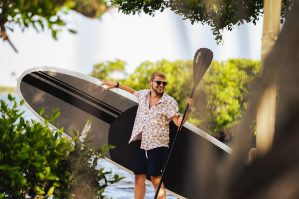 A man in sunglasses and a floral shirt stands holding a large paddleboard and paddle, surrounded by greenery near the water, smiling cheerfully.