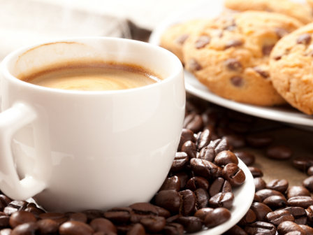 A white cup filled with coffee surrounded by coffee beans sits next to a plate of chocolate chip cookies on a table.