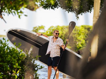 A man wearing a floral shirt and sunglasses holds a paddle while standing next to a paddleboard by a water body, surrounded by lush greenery.