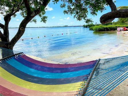 A colorful hammock is stretched between two trees on a sandy beach, overlooking calm, clear water with a distant shoreline and boats.