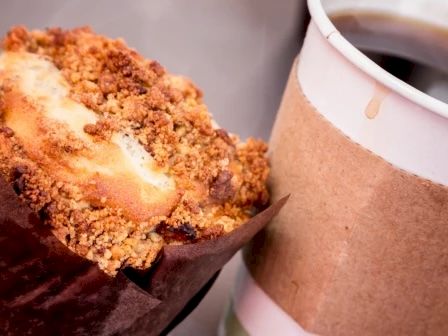 A close-up of a coffee cup with a cardboard sleeve and a crumbly muffin in a paper wrapper is shown. The coffee has a small spill on the sleeve.