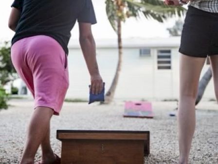 Two people play cornhole outdoors. One is throwing a bean bag, and the other holds a drink. Palms and a building are in the background.