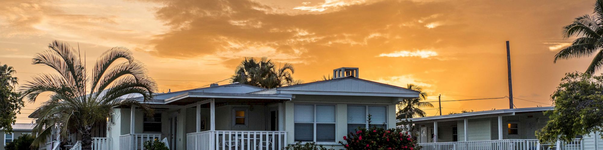 A serene waterfront scene depicts a cozy house with a boat docked nearby, framed by palm trees and bathed in a warm, glowing sunset.