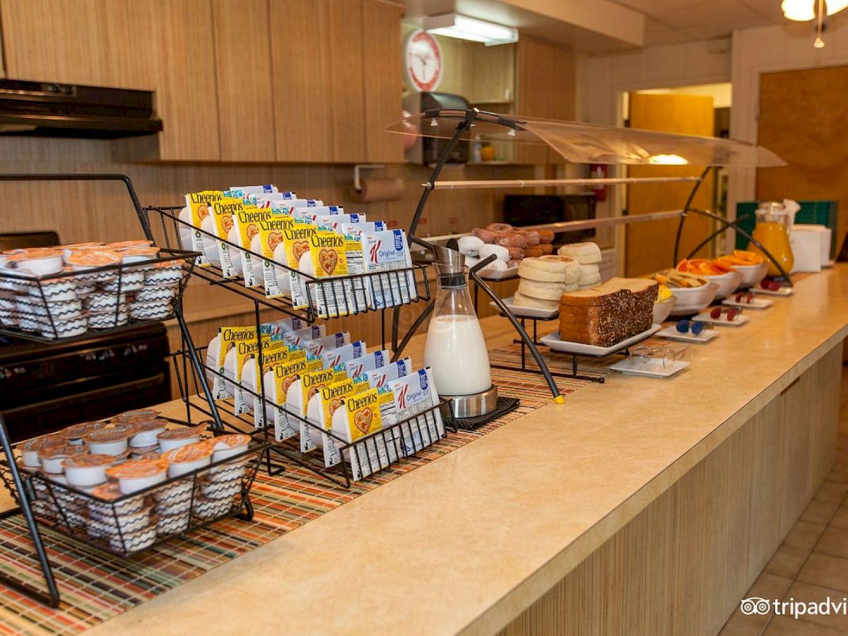 The image shows a breakfast buffet with cereal, milk, muffins, bread, bagels, fruits, and juice cartons on a counter in a kitchen setting.