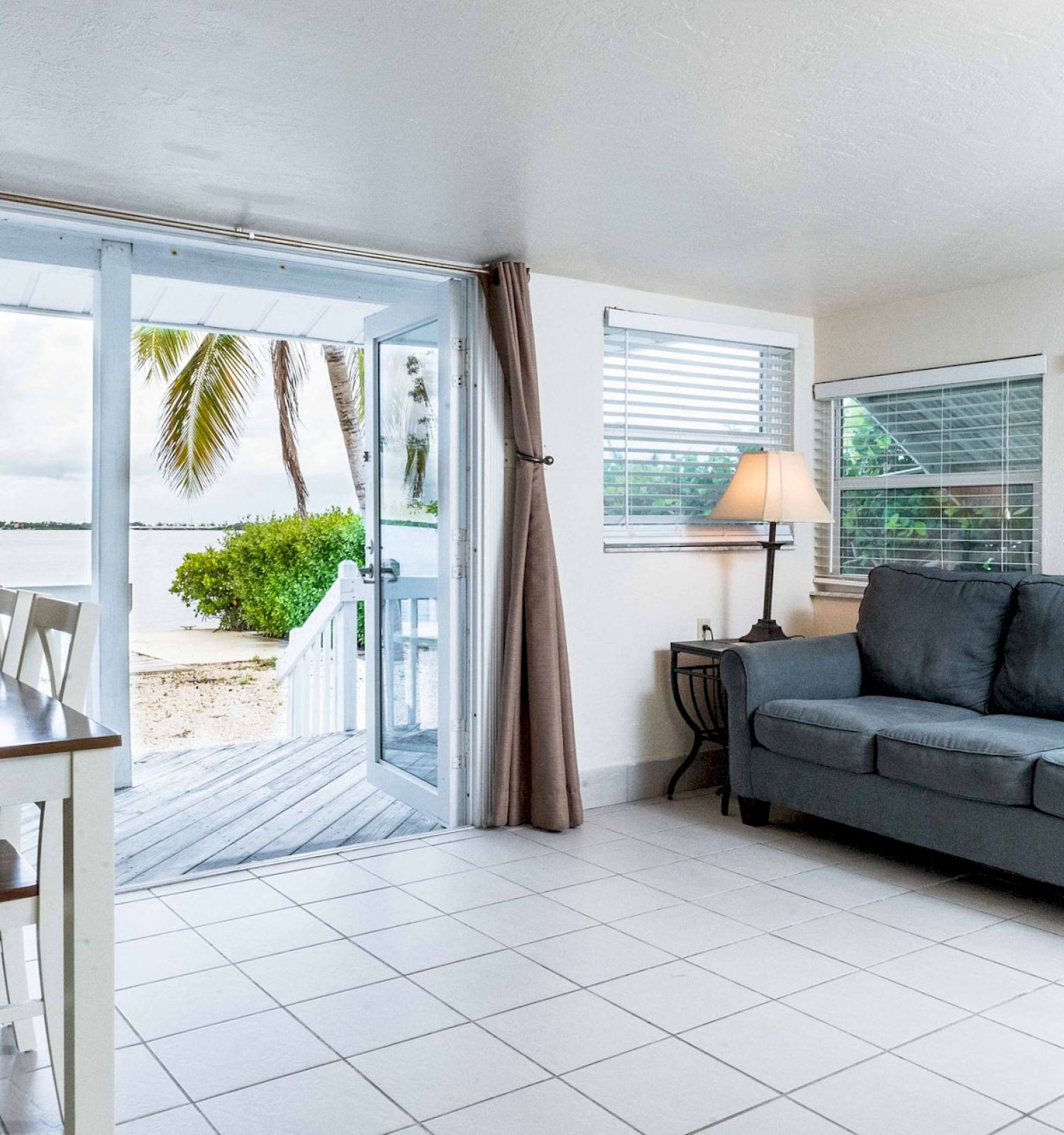 A bright living room with a grey couch, small dining table, and glass doors opening to a patio with a beach view. Pale floor tiles are seen.