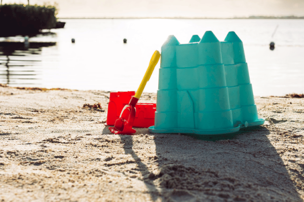 A blue sandcastle mold, a yellow shovel, and a red bucket on a sandy beach near calm water with a shoreline and distant background.