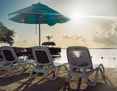 Beach scene with lounge chairs, umbrellas, and a calm body of water under a partly cloudy sky during sunset.