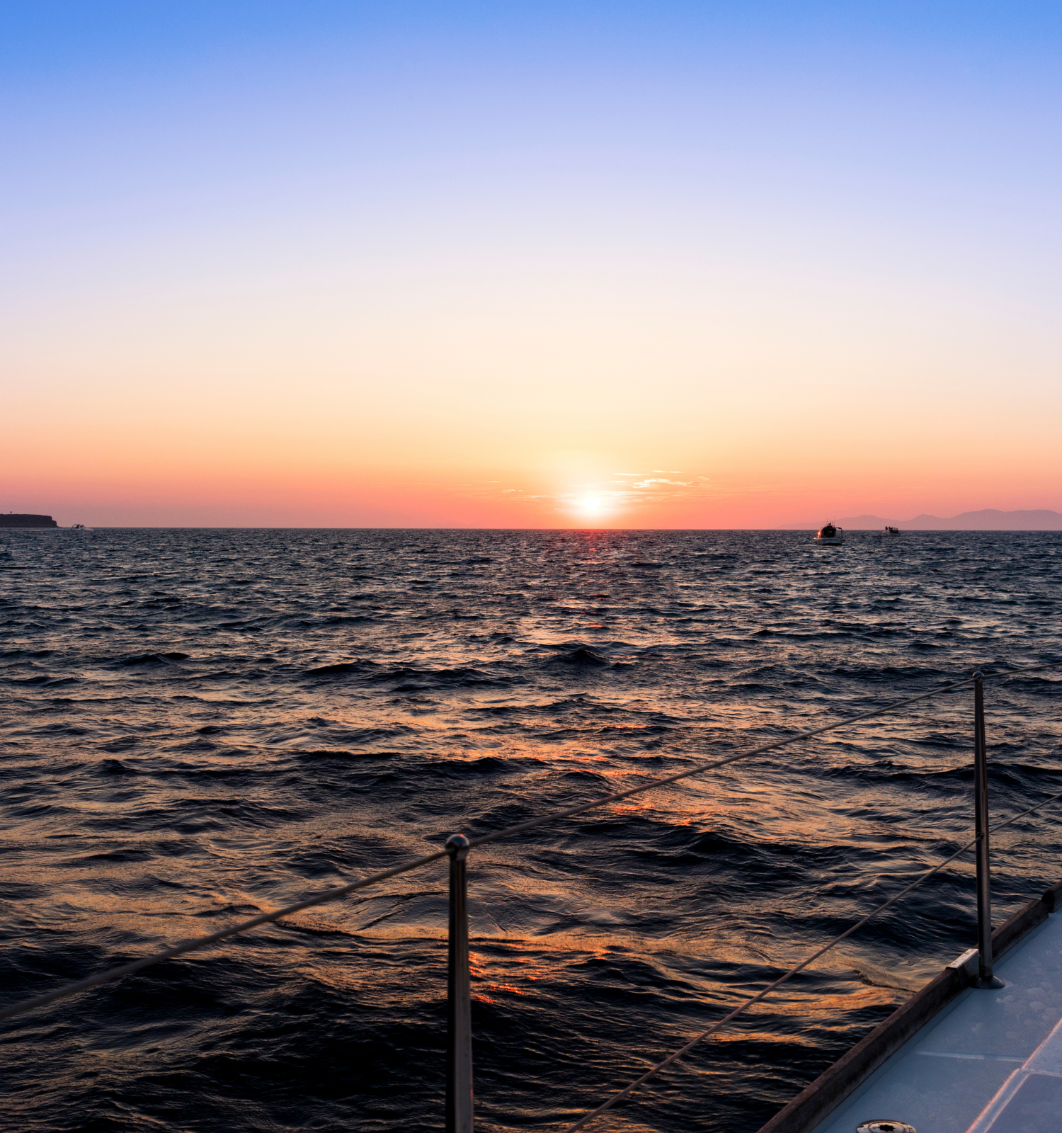 A boat is sailing toward a serene sunset over a calm, vast ocean, with the horizon meeting the colorful sky in the background.