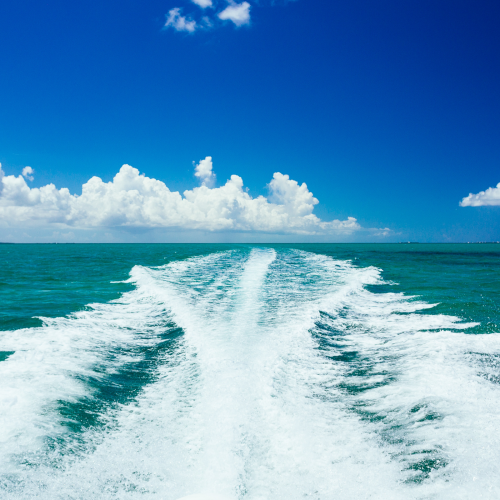 This image shows the wake of a boat on a large body of water under a clear blue sky with some clouds.