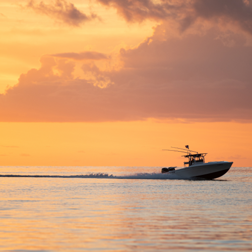 A boat cruises across calm waters during a vibrant sunset, with orange and pink hues illuminating the sky and clouds, creating a serene scene.