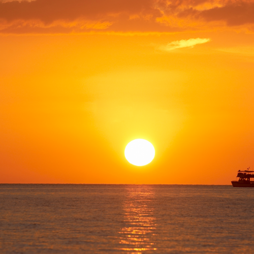 A stunning sunset over calm ocean waters, with a small boat silhouetted against the glowing orange sky and sun.