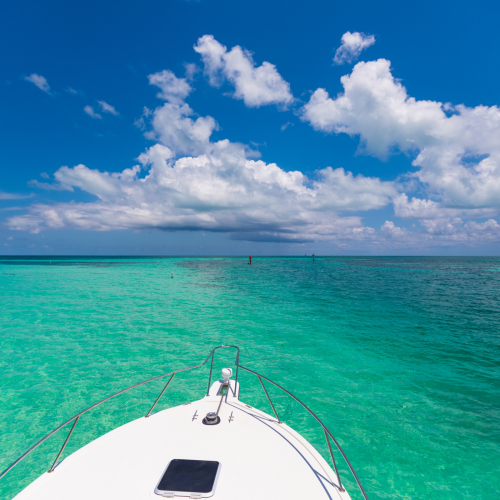 The image shows the front of a boat sailing on clear turquoise water under a bright blue sky with fluffy white clouds.