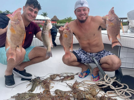 Two people are on a boat showing off their catch, including fish and lobsters, while smiling for the camera.