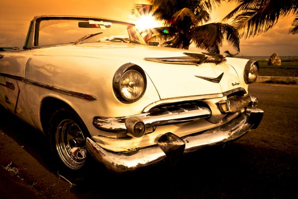 A vintage white convertible car parked near a beach with palm trees at sunset, creating a nostalgic and serene scene.