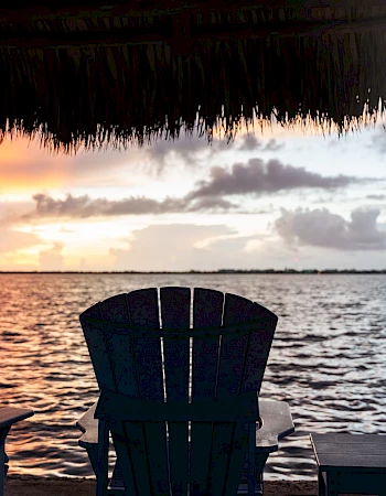 Three chairs facing a tranquil body of water at sunset, with vibrant sky colors and a wooden pier extending into the distance.