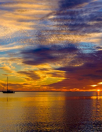 A serene sunset over calm waters with vibrant clouds and a distant sailboat silhouetted against the horizon, creating a tranquil scene.