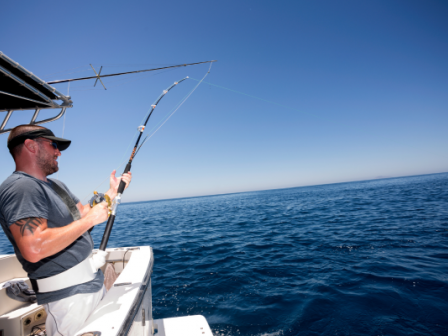 A person stands on a boat holding a fishing rod, engaged in deep-sea fishing under a clear blue sky.