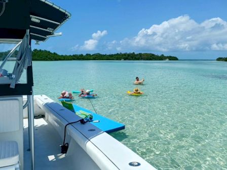 A boat is anchored in clear, shallow water with people floating nearby. It's a sunny day with a view of distant green islands.