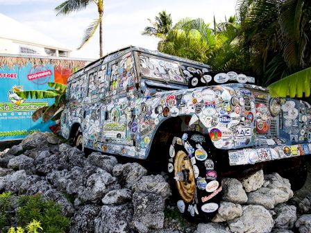 A vintage SUV covered in stickers is displayed on rocks, with tropical plants and a colorful sign in the background.