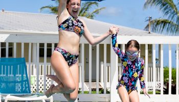 Two people in swimsuits are joyfully jumping into a pool, holding hands, with blue umbrellas and palm trees in the background.