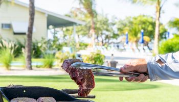 A person is grilling seasoned steaks outdoors with tongs, in a sunny garden with trees and a building in the background.