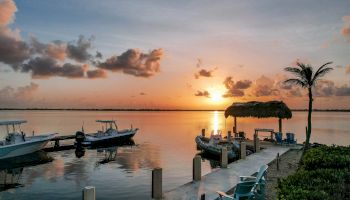 A serene sunset over a calm lake, with boats docked beside a small pier and a lone palm tree on the right.