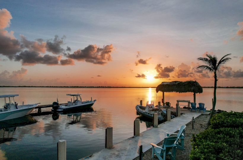 A serene sunset over a calm lake, with boats docked beside a small pier and a lone palm tree on the right.