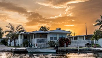 A cozy waterside house with a balcony and boat docked, surrounded by palm trees and a scenic sunset sky reflecting on the water's surface.