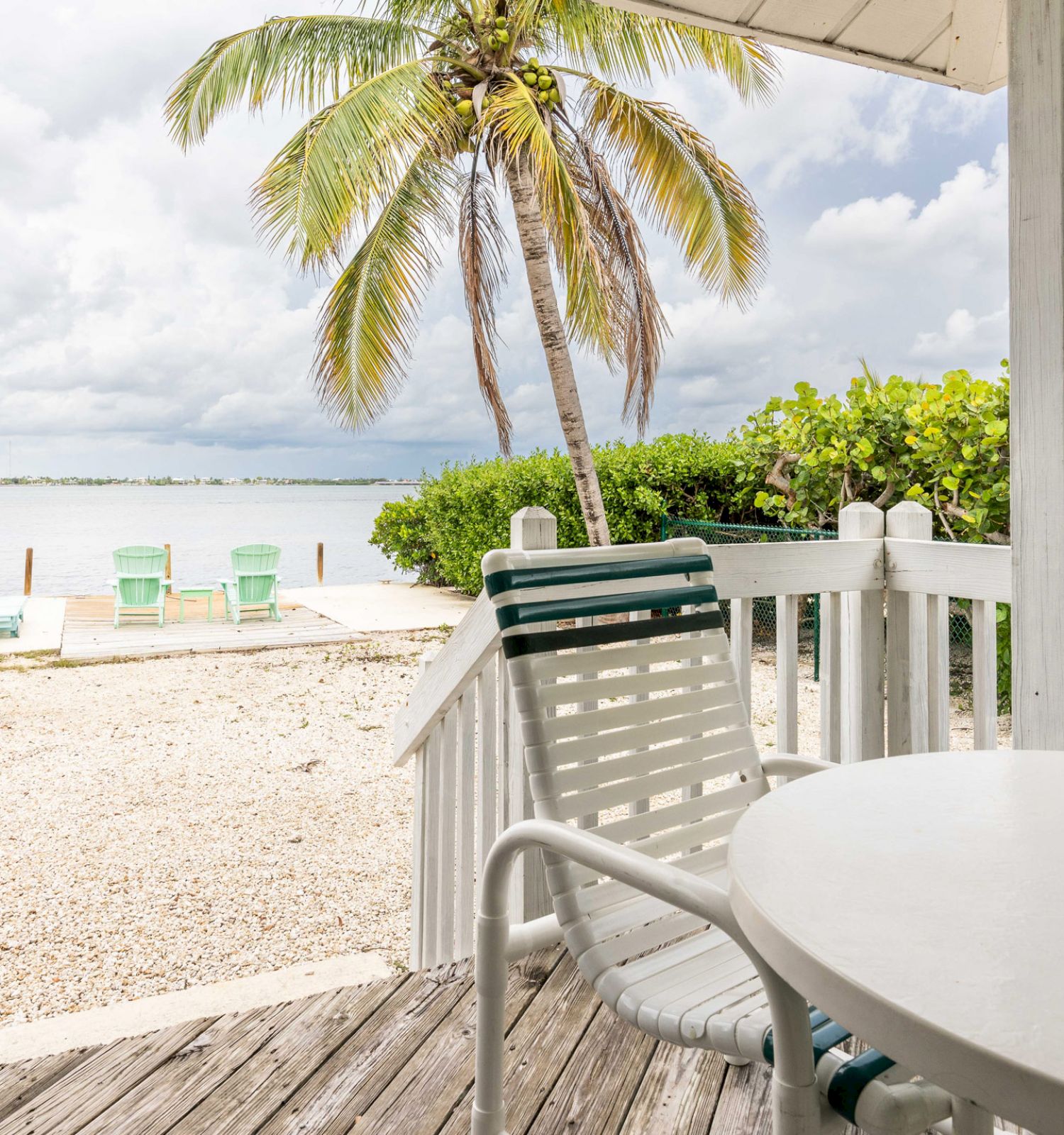 A beach view from a porch with a white table and chairs, overlooking a sandy shore, green lawn chairs, a palm tree, and the ocean under a cloudy sky.