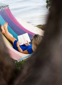 A person is relaxing in a colorful hammock by a body of water, reading a book. The scene is partially framed by tree branches, creating a serene ambiance.