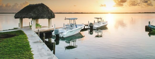 A serene waterfront scene at sunset features a thatched-roof hut, docked boats, calm water, and partly cloudy skies with brilliant sunlight.