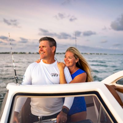 A smiling couple is enjoying a boat ride on the water during a beautiful, cloudy sunset. The woman embraces the man as they look towards the horizon.