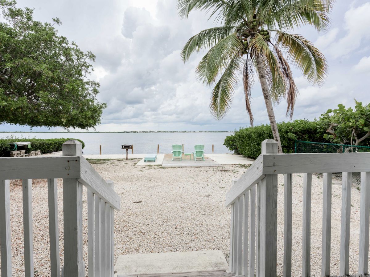 A view from a porch steps leading to a serene lakeside area with lounge chairs, a grill, palm tree, and cloudy sky above the tranquil water.