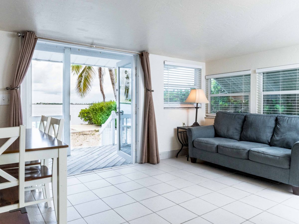 A bright, airy living space with a grey sofa, a dining table, and a view of a tropical beach through sliding glass doors.