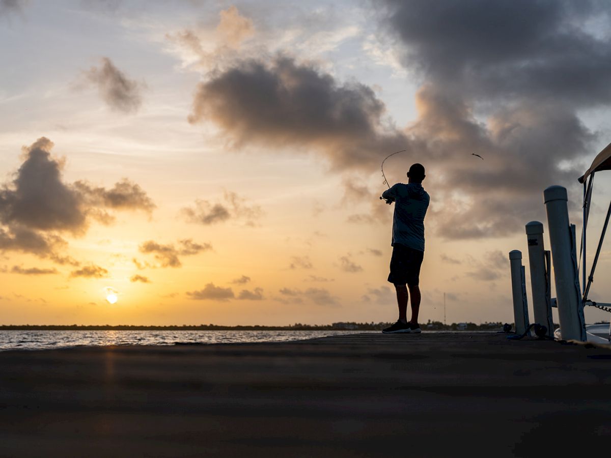 Silhouette of a person fishing on a dock at sunset, with clouds and calm water in the background.