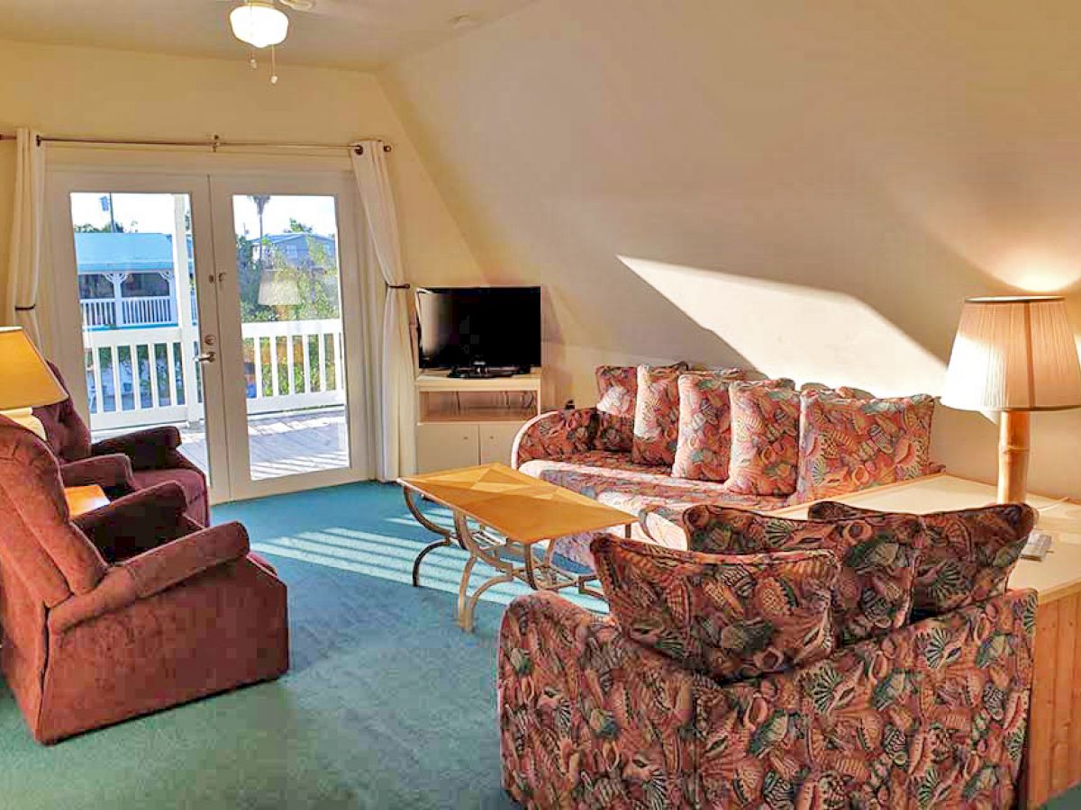 A cozy living room with floral-patterned furniture, a TV, and sliding glass doors leading to a balcony. Two lamps and a coffee table are also visible.