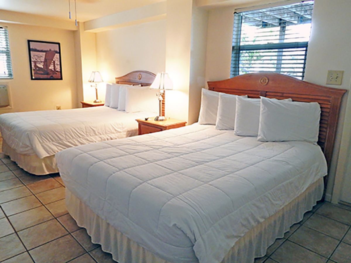 The image shows a hotel room with two double beds, white linens, bedside tables with lamps, wall art, tile floor, and windows with blinds ending the sentence.