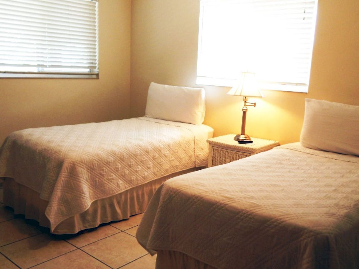 This image shows a bedroom with two twin beds, a bedside table between them with a lamp, and two windows with blinds.